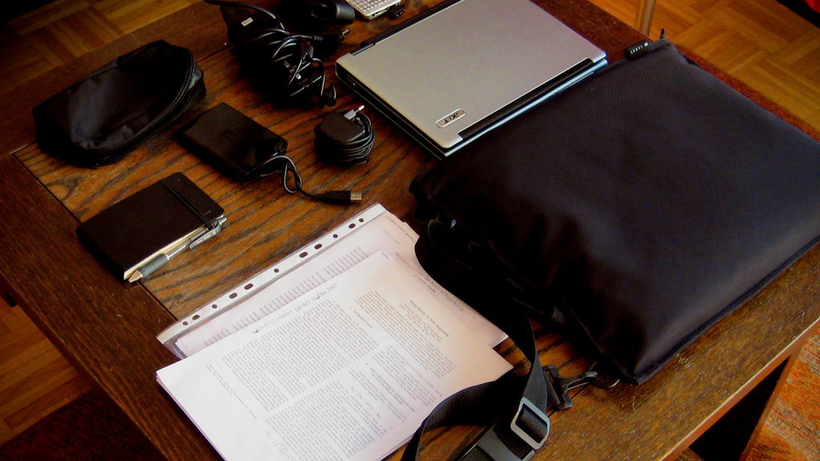 A wooden desk with a closed laptop, a laptop bag, chargers, a notebook and printed papers