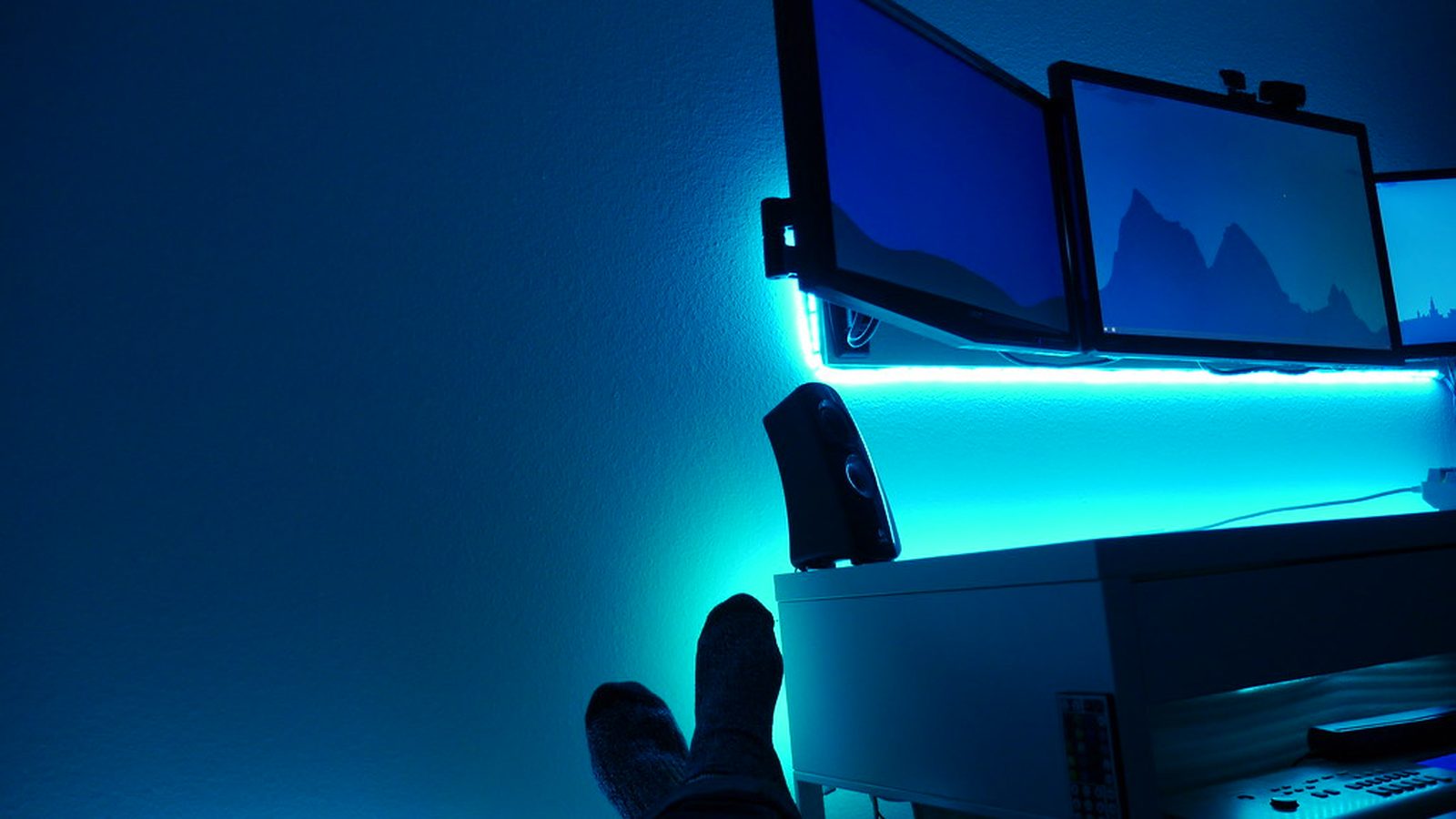 A desk lit blue at night, with three monitors and feet up on the desk